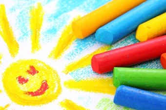 drawing of a sun with with a smily face and blue sky and green grass with various colors of crayons next to it