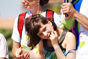 Young Girl displaying Rett Syndrome features looks at the camera while standing by adults