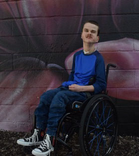Young man in a wheelchair with a tracheotomy in front of a mural